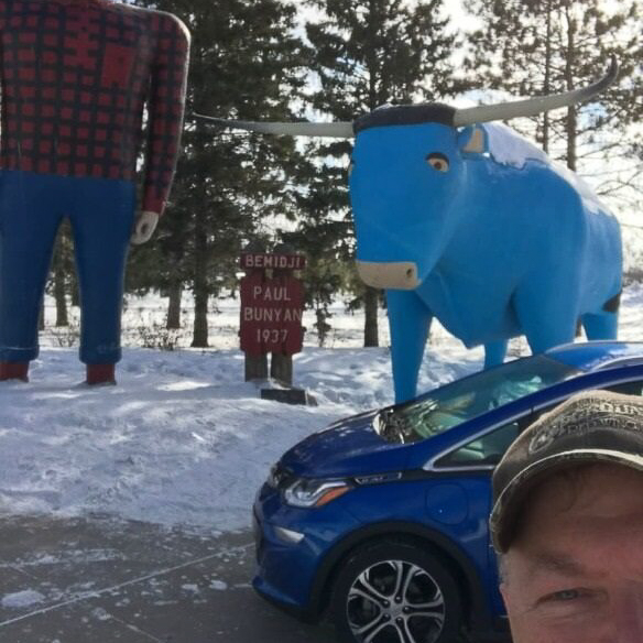Man taking a selfie with a blue electric vehicle and a monument in a park