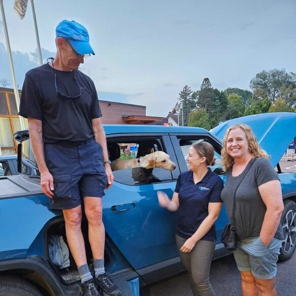 group of 3 outside of a blue electric truck with a dog in the back seat