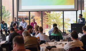 Four people siting at the front of the room during a panel as a presenter stands off to the side.